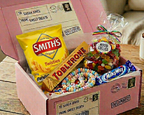 A pink subscription snack box filled with assorted candy and sweets, including a bag of Smith's Twists, a Toblerone bar, and a bag of Milky Way, displayed on a wooden table next to a mug.