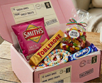 A pink subscription-style candy box filled with assorted sweets, shown open on a wooden table with a mug nearby. Visible candy brands include Smith's, Toblerone, Milky Way, and M&M's.