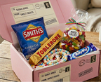 A pink personalized candy gift box filled with assorted sweets, placed on a wooden table next to a mug. Visible candy brands include Smith's, Toblerone, Milky Way, and Mints of Lollars.