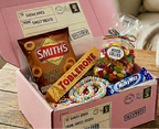 A pink subscription sweet treat box filled with assorted candy and snacks, including a Smith's Burger Bites bag, a Toblerone bar, a Milky Way candy bar, and a bag of mixed lollipops, displayed on a wooden table next to a mug.